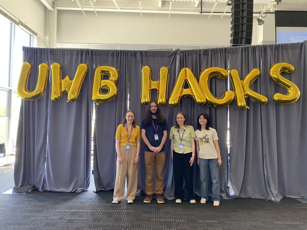 Three girls and one guy standing in front of gray curtains and golden bubble letter balloons reading 'UWB Hacks.'
