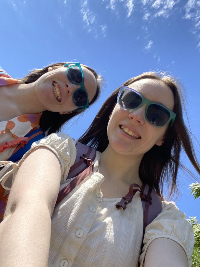 Two girls wearing sunglasses in front of a bright blue sky with fluffy white clouds.