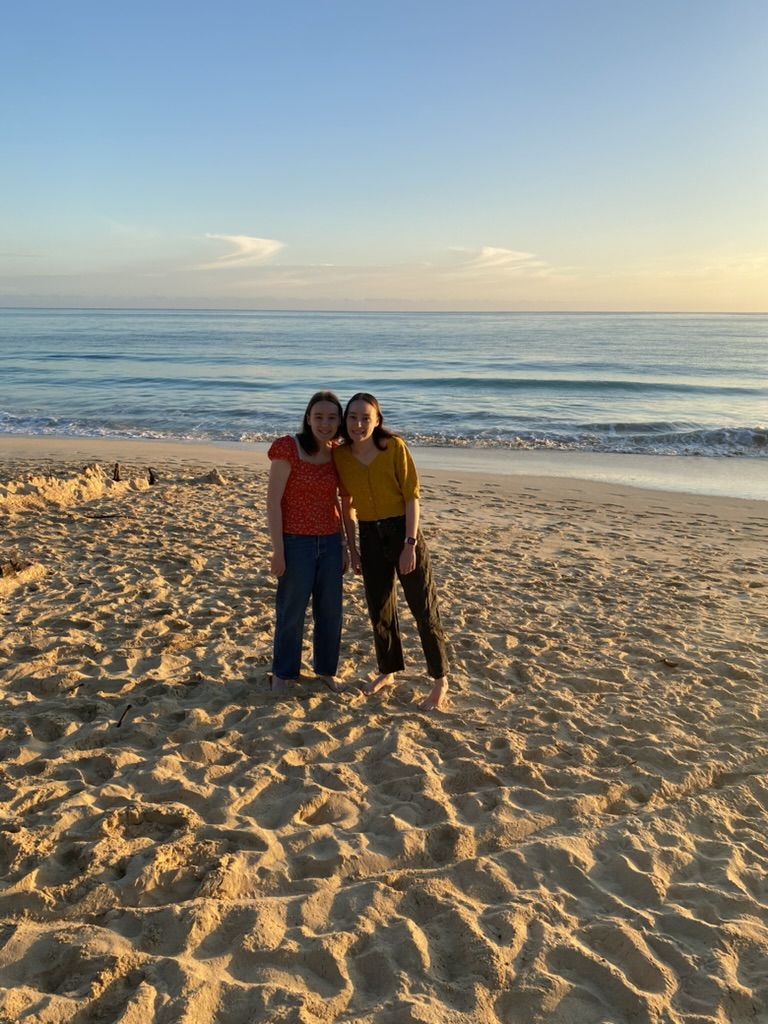 Two girls standing on a sandy beach at sunrise.