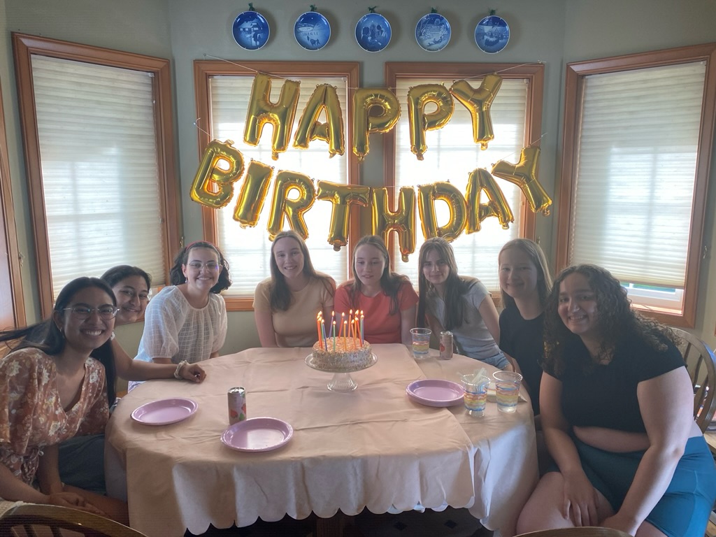 Eight girls sitting in front of happy birthday balloons at a pink table with a pink cake.