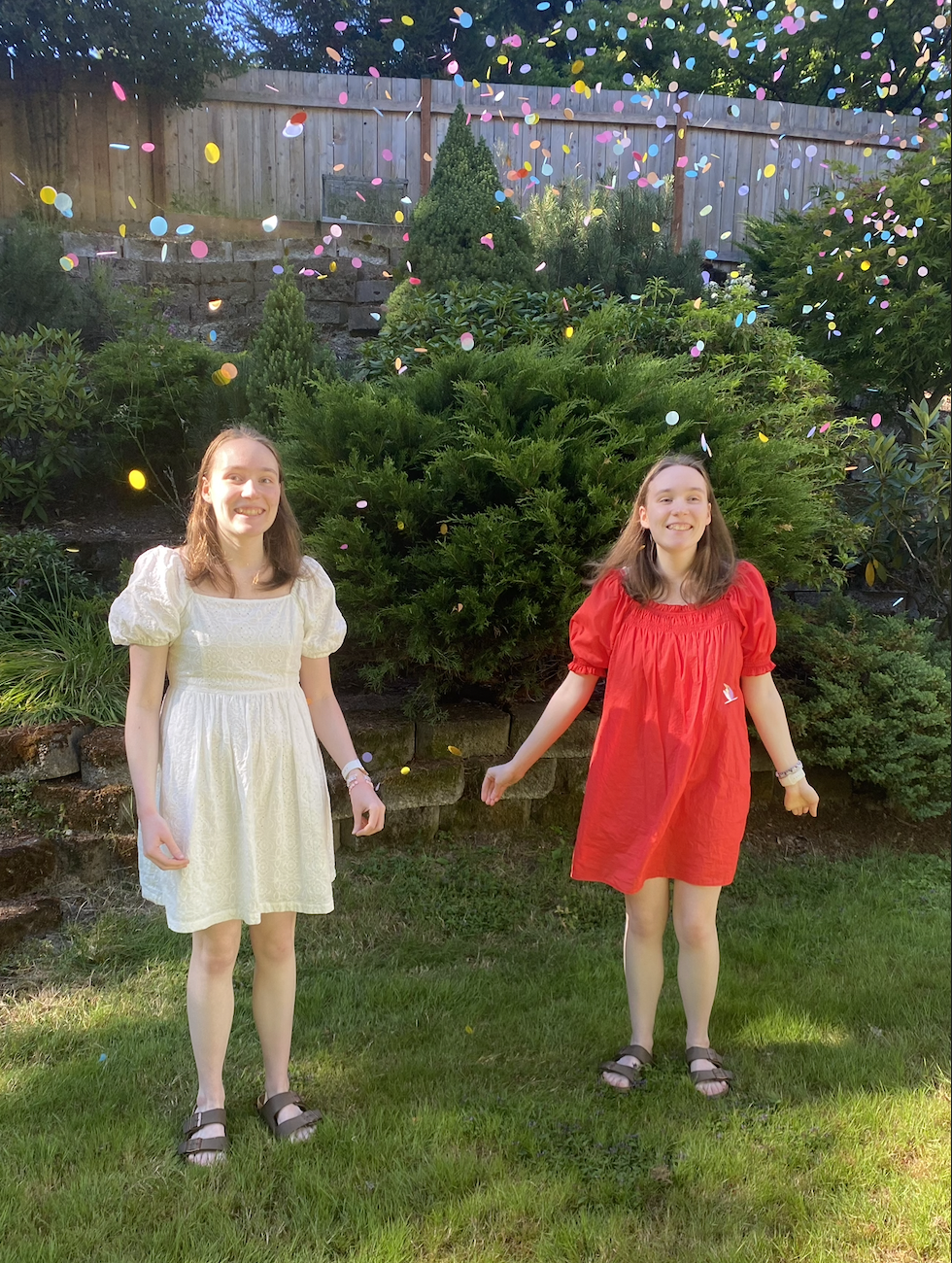 Two girls wearing dresses throwing rainbow confetti in the air in front of a hill of plants.