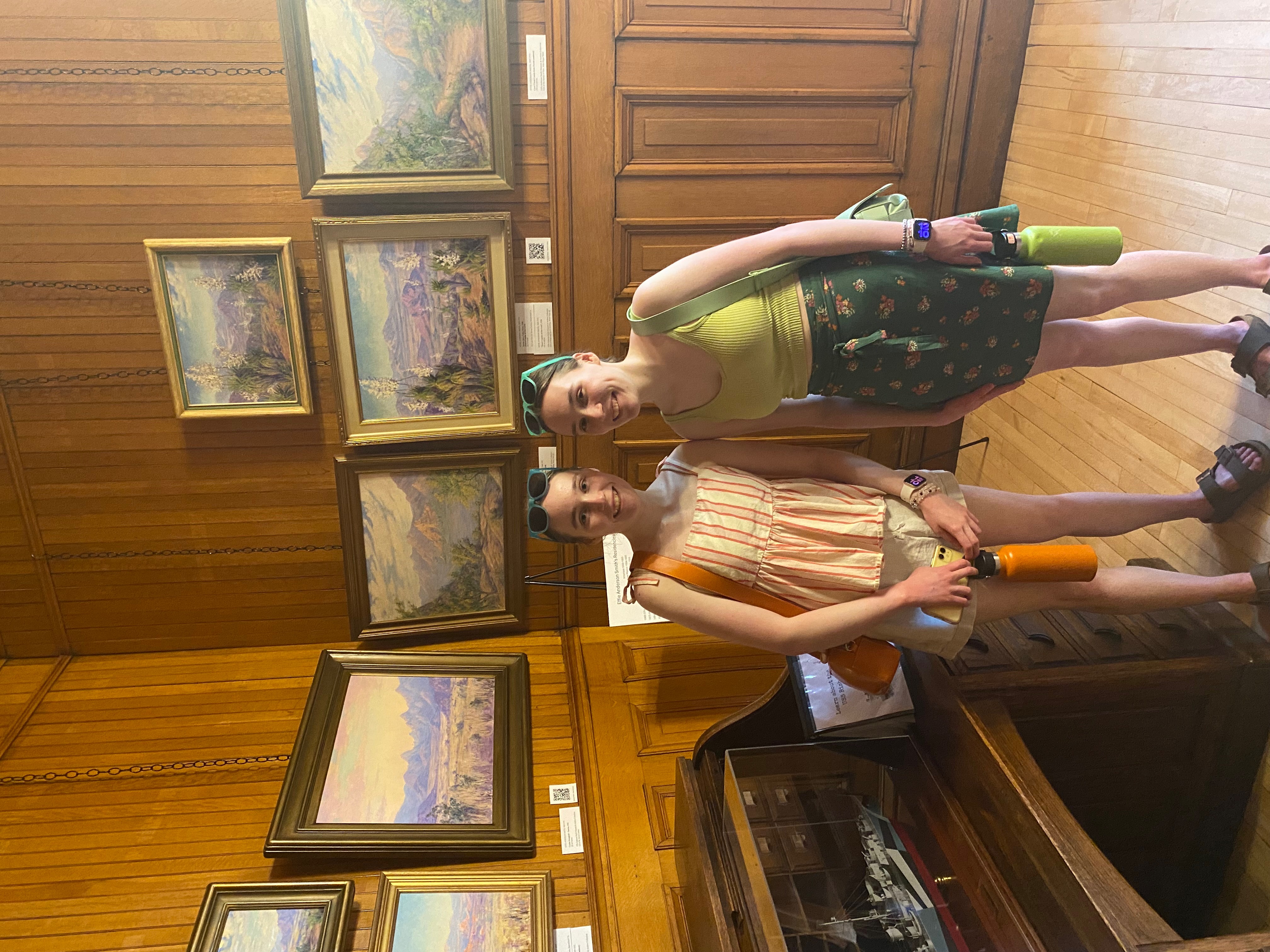 Two girls standing in front of desert scenery paintings in a museum.