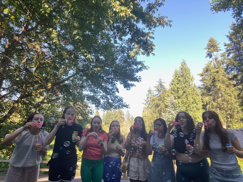 Eight girls blowing bubbles outside surrounded by lots of green trees.