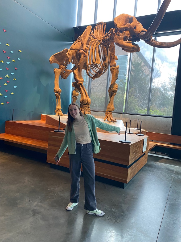 A girl with her arms in the air in front of a woolly mammoth fossil.