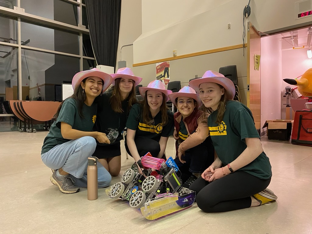 Five girls in pink cowboy hats crouched around a robot on the floor.