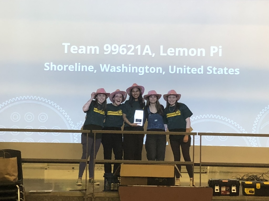 Five girls in pink cowboy hats holding a trophy in front of a screen projector.