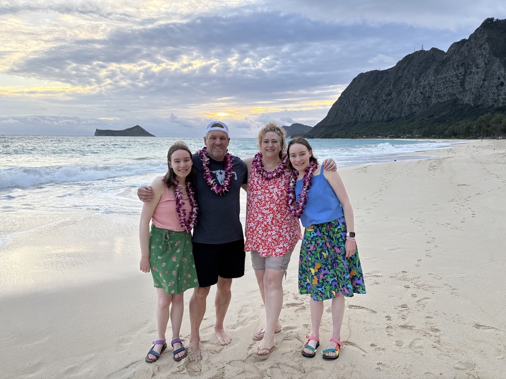 A family of four standing on a sandy beach at sunrise wearing leis.