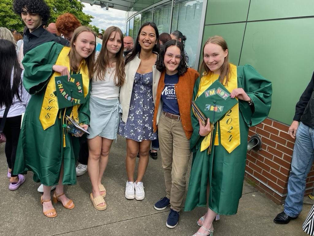 Five girls standing smiling, two wearing graduation gowns and holding up their graduation caps.