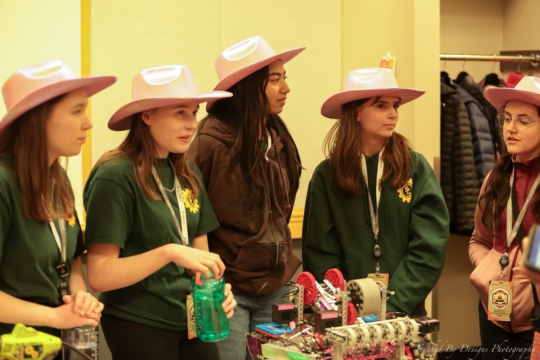 Five girls in pink cowboy hats talking and standing around a table with a pink and purple robot.
