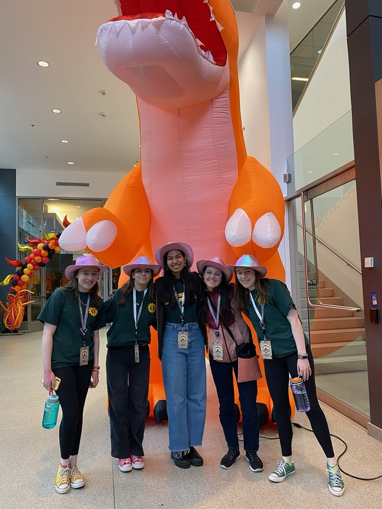 Five girls in pink cowboy hats standing in front of a blow up orange dinosaur.