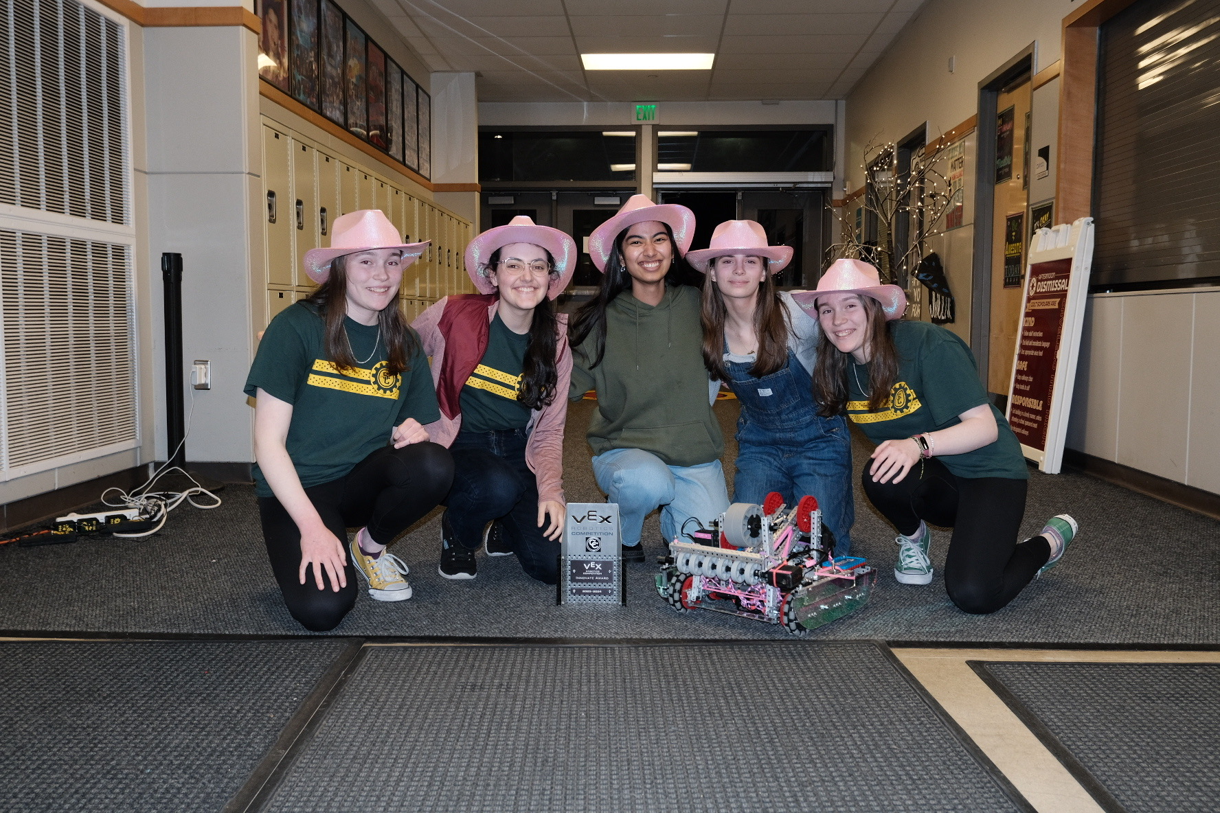 Five girls in pink cowboy hats crouched on the floor around a pink robot and trophy.
