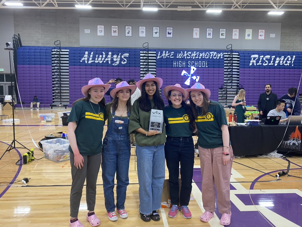 Five girls in pink cowboy hats standing in front of purple bleachers holding a trophy.