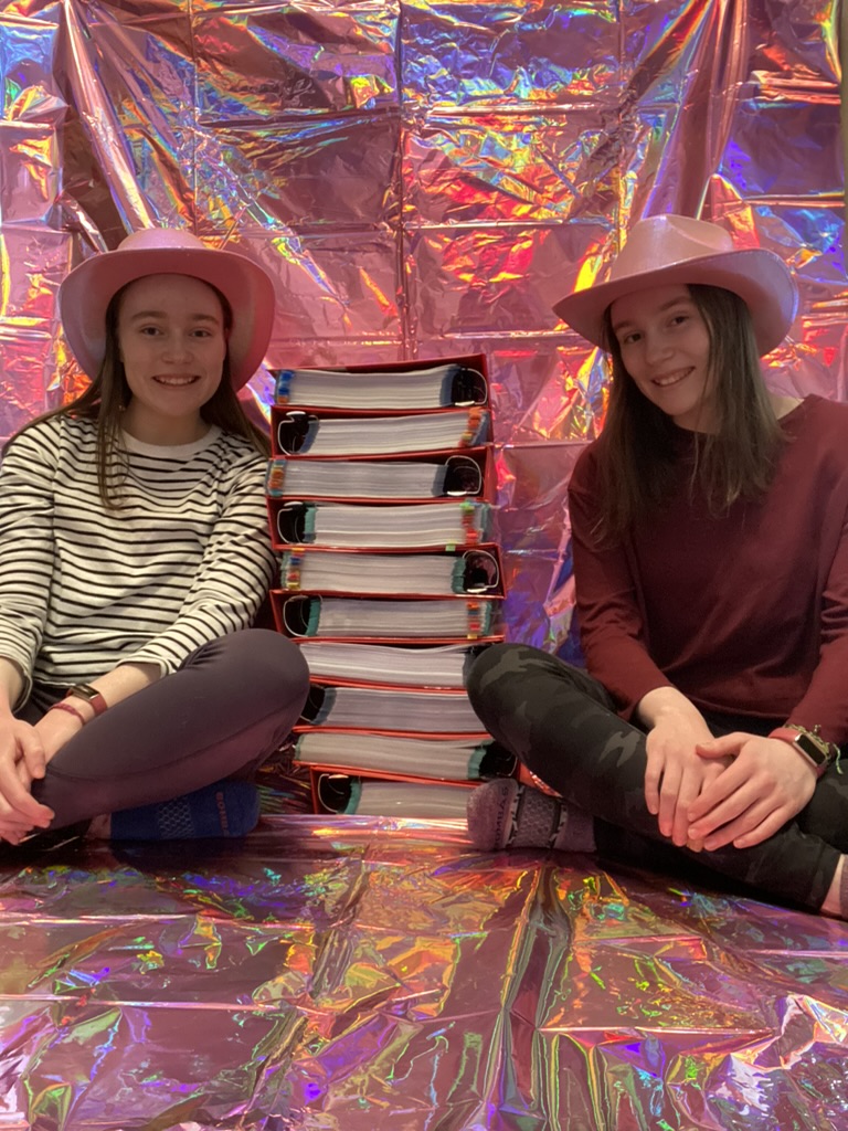 Two girls in pink cowboy hats sitting in front of a pink sparkly wall with ten big red binders stacked up between them.