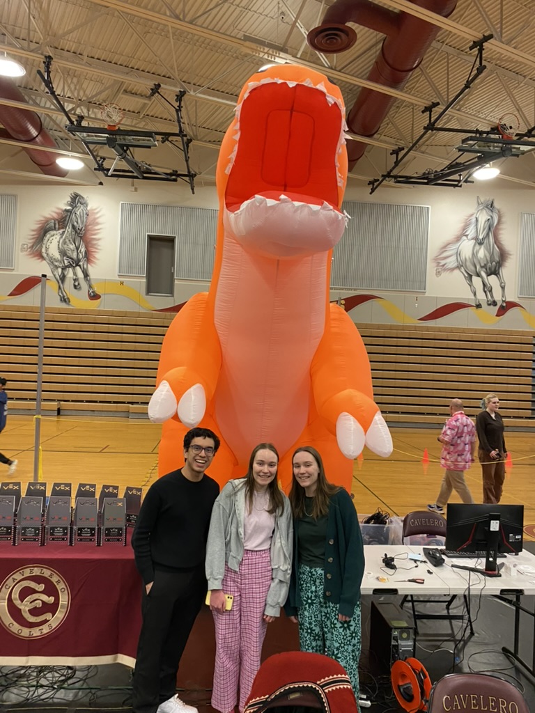 Two girls and one guy standing in front of large, orange inflatable dinosaur.