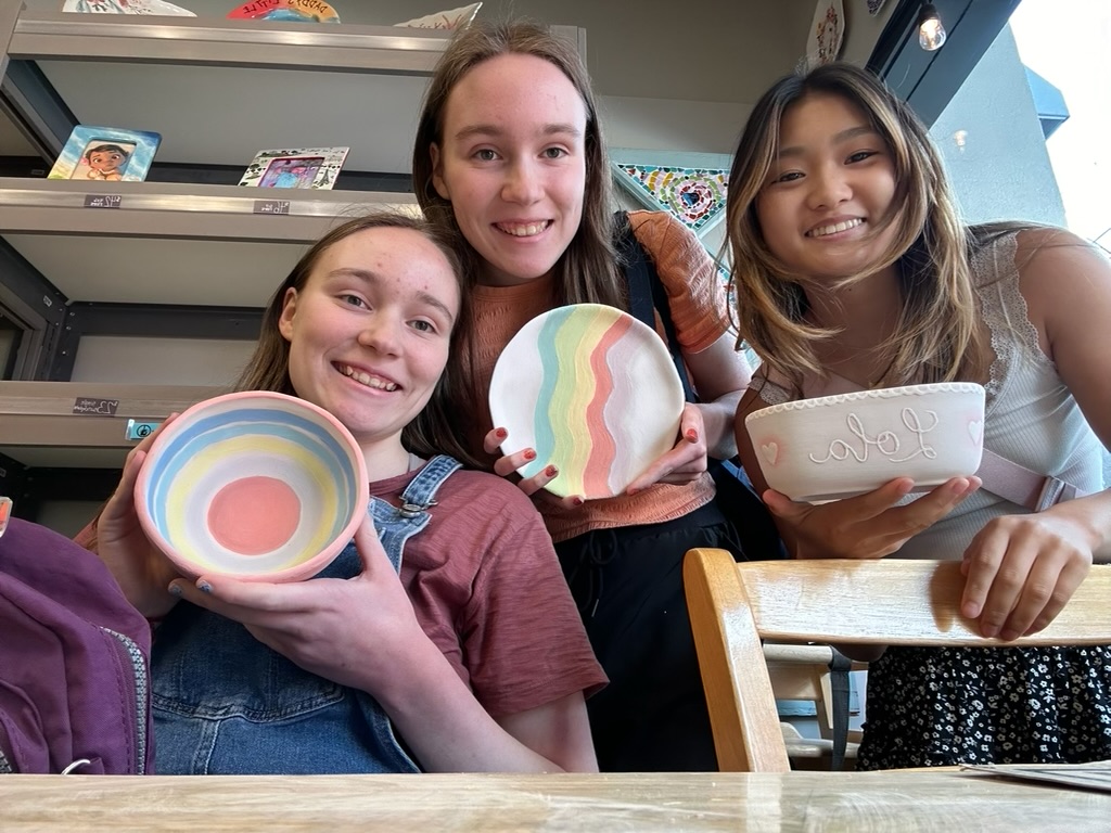 Three girls holding painted ceramic pieces around a wooden table.