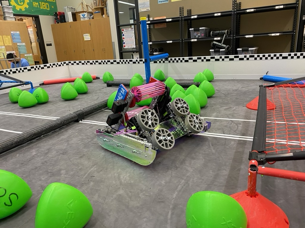 Pink and purple robot sitting in a robot field with red and blue netted goals and green acorn shaped game elements.