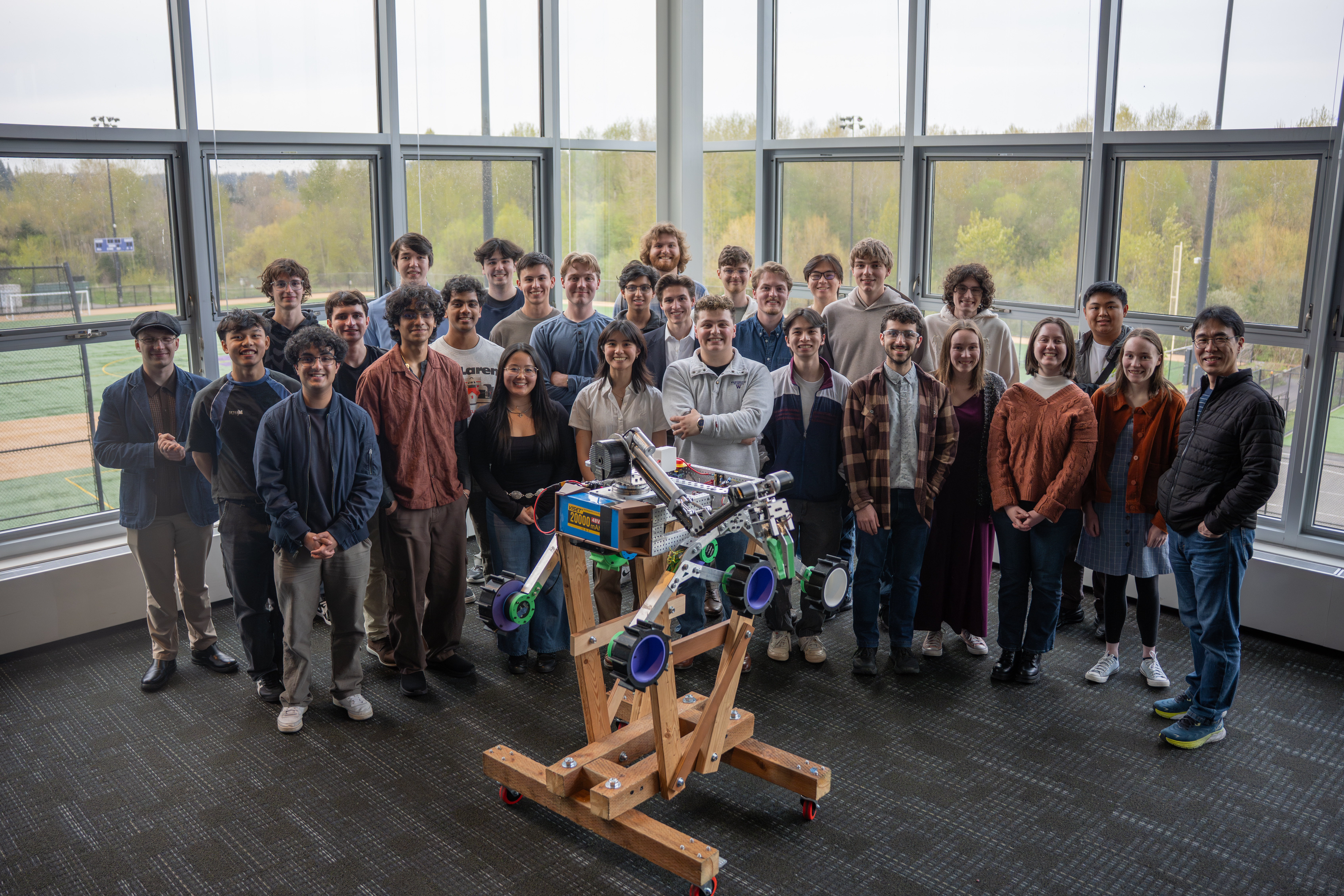 A group of students standing in front of big windows beside a rover on a wooden stand.