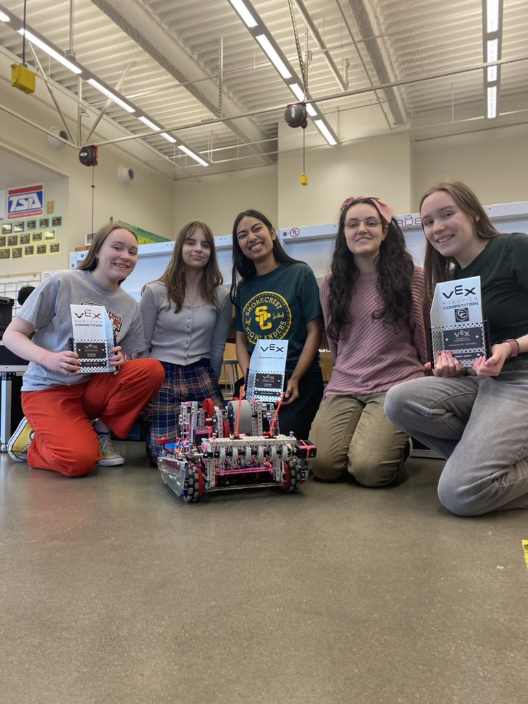Five girls holding three trophies crouched around a robot on the floor of a classroom.