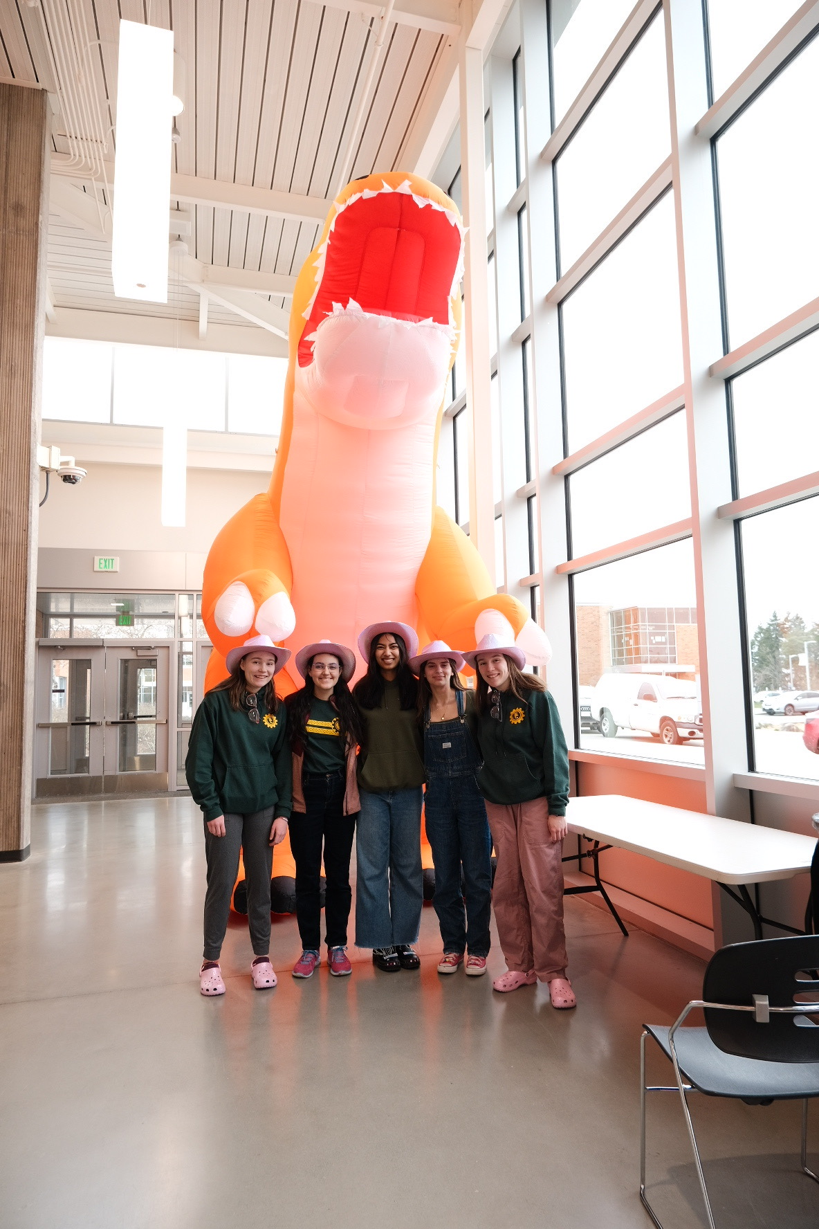 Five girls in pink cowboy hats standing in front of a blow up orange dinosaur next to big windows.