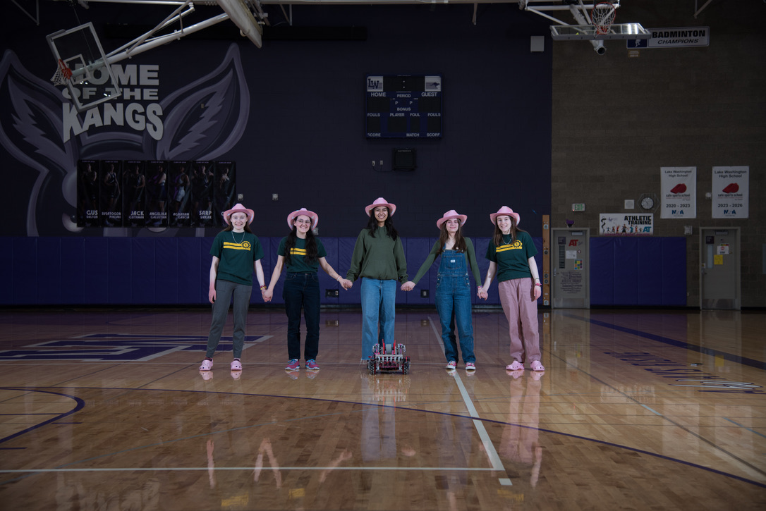 Five girls in pink cowboy hats holding hands with a robot at their feet in a gymnasium with purple walls.