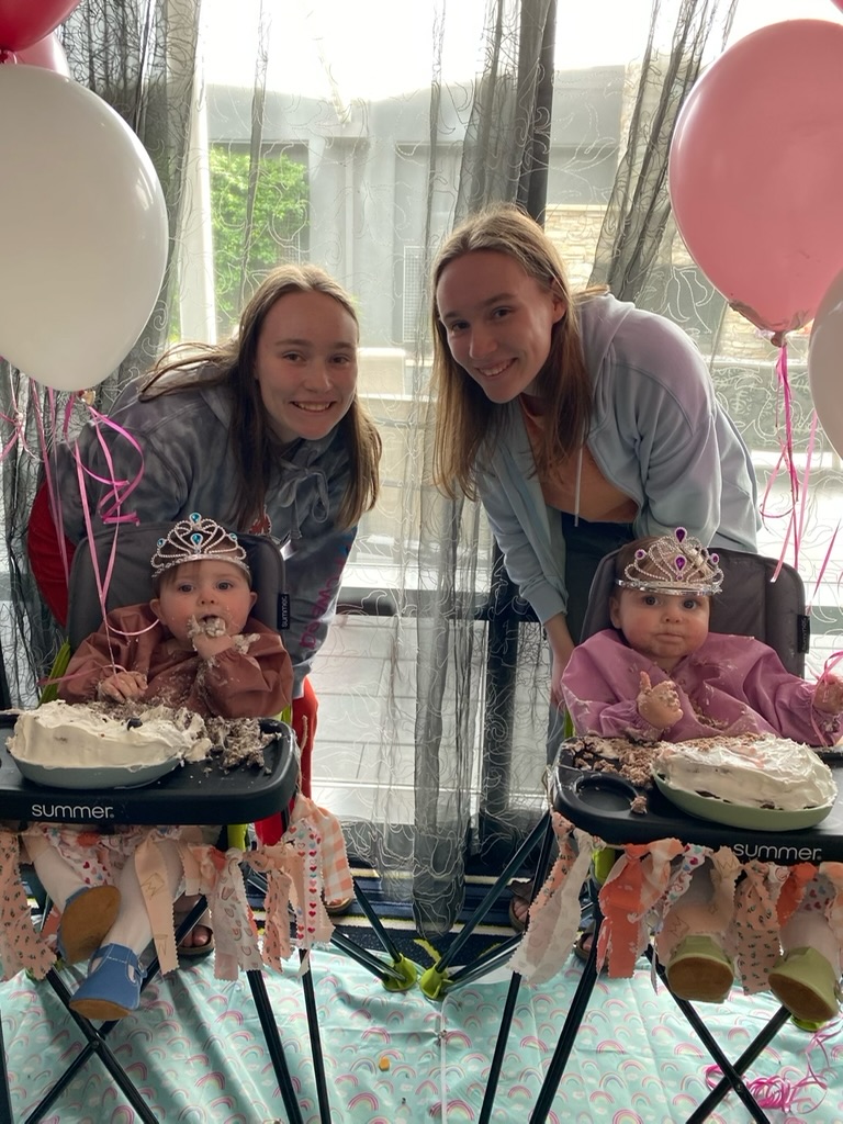 A set of older twins smiling beside baby twins who are eating cake and wearing tiaras.